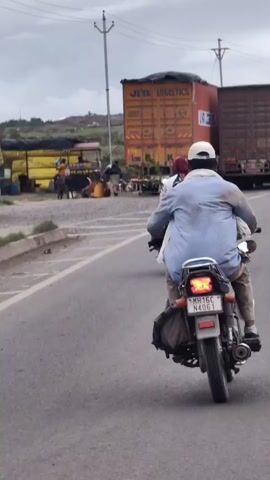 This video shows two people riding on a motorcycle on a road. The person in front is wearing a blue shirt and a white cap, and is holding what appears to be a newspaper. The person behind is wearing a green saree and a red headscarf, and is holding a bag. They are riding on the right side of the road, and there are large trucks on the left side of the road, possibly parked. The road appears to be paved and is likely in a rural or suburban area, given the presence of the trucks and the landscape in the background. The sky is overcast.