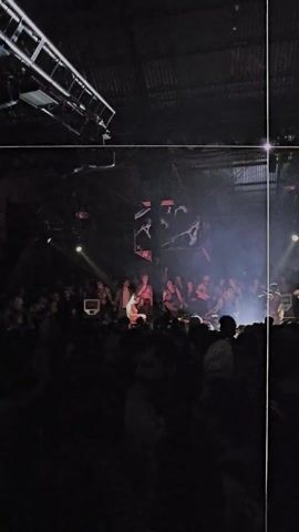Here is a description of the video:
The video shows a crowded dance floor at a nightclub. There is a DJ playing music on a platform in the center of the room. The crowd is diverse, with many people dancing, and some are partially undressed. The lighting is low and dramatic, with spotlights shining on the DJ and the crowd. There is a large cube hanging above the DJ with abstract images projected on it. The building appears to be an old industrial space with a corrugated metal roof and exposed beams.
