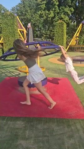This is a video of a playground on a sunny day. In the foreground, a purple and yellow merry-go-round is spinning on a red mat. A young girl with blonde hair is hanging from the merry-go-round with her hands, her legs extended in front of her. Another person, possibly an adult, is on the left side of the merry-go-round, also holding onto it. In the background, there are tall green hedges, trees, and yellow playground equipment. The grass around the red mat is green. There are other children visible on some of the background equipment. The video pans and rotates with the merry-go-round.