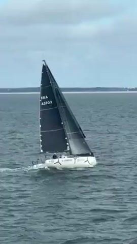 The video shows a white sailboat with a black sail moving on the water. The sail has the text "FRA 43933" in white. The boat also has the words "VL ENERGIES" written on it. The water is a dark shade of blue, and the sky is overcast with a light blue hue. A distant shoreline is visible in the background.