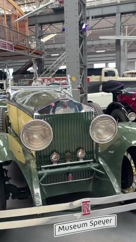 The video shows a panning shot of a museum exhibit featuring vintage vehicles and aircraft. The camera begins on the front of a dark green and yellow Rolls-Royce convertible, showcasing its iconic grille, headlights, and the "Museum Speyer" sign on its bumper. As the camera pans to the right, it reveals a red convertible Rolls-Royce with a spare tire on the side and wooden running boards. Further to the right, a large, dark red vintage bus or carriage is visible. In the background, various aircraft are suspended from the ceiling, including a sleek silver jet and a yellow and red biplane. The overall environment appears to be a large, well-lit indoor exhibition hall with exposed metal beams and industrial architecture. The video is silent.