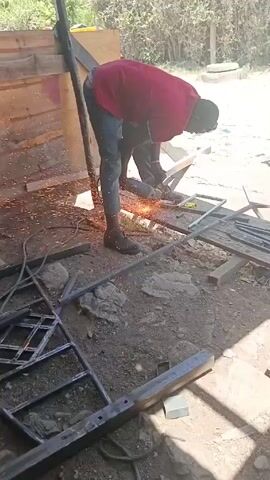 The video shows a man cutting metal with a power tool. The man is wearing a red shirt, gray pants, and dark shoes. He is standing in a cluttered outdoor workspace, with metal pieces and tools scattered around. He is leaning over a small table, where he is cutting a piece of metal with an angle grinder. Sparks are flying from the cutting point. A metal structure is visible in the foreground, possibly part of a gate or fence. A wooden structure, possibly a shed, stands in the background, along with greenery. The scene is well-lit, but the bright light makes it difficult to see details clearly.