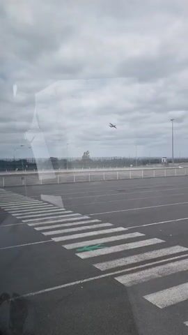 A video filmed from inside a vehicle shows a crosswalk in an empty asphalt parking lot under an overcast sky. A plane descends, flying from right to left in the background, eventually passing out of view. Several tall light poles and a fence are visible in the background, along with a few parked trucks. A green marking is visible on the asphalt in the middle of the crosswalk. There are no people visible.