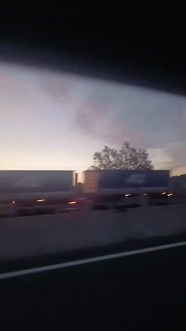 The video shows a view from a vehicle traveling on a highway at dusk. The sky is a mix of blue and pink hues, with some clouds. The landscape consists of trees and hills. A white truck is seen driving on the highway.