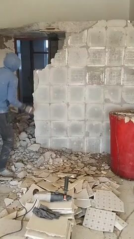 A person wearing a blue hooded sweatshirt and dark pants is demolishing a tiled wall with a hammer. The wall is made of large square tiles with a grid pattern and has a large hole at the top left. Debris is scattered across the floor, including tile fragments and a power drill. A red cylindrical object is visible to the right of the wall. The room is mostly empty and poorly lit. There is a doorway in the background. The person is striking the wall with the hammer repeatedly, causing more tiles to break and fall to the floor.
