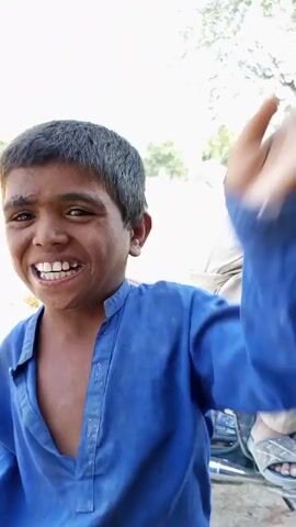 A young boy with dark hair and skin smiles at the camera, wearing a blue tunic. His right hand is raised in a waving gesture. The background is out of focus, showing some greenery and a light-colored sky. The boy appears to be outside. The video consists of a single shot that lasts for about four seconds.