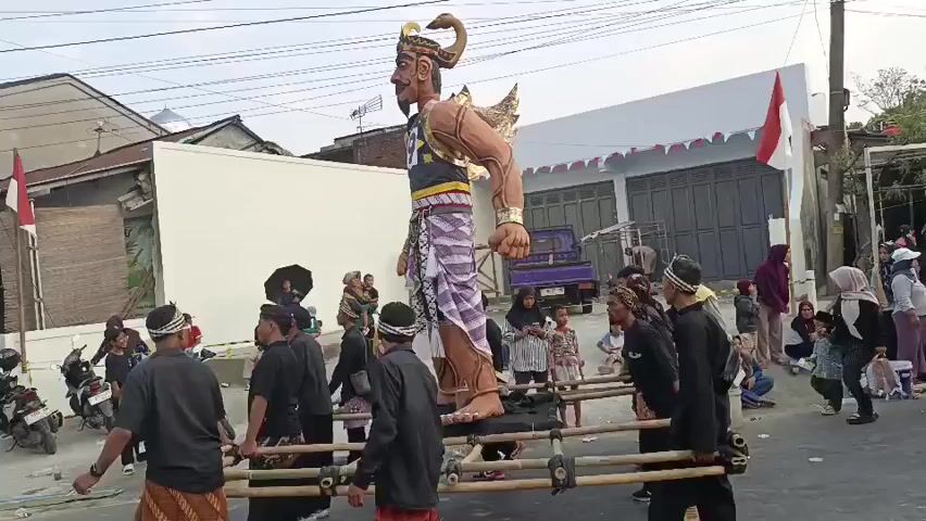 A large, colorful effigy of a male figure with a horned headdress and elaborate facial features is being carried on a wooden platform by a group of men. The effigy is dressed in a striped sarong and a dark top with gold trim. The men carrying it are wearing dark clothing and traditional head coverings. They appear to be participating in a procession or parade, as there are onlookers on the sides of the street, including women and children. Some people are holding umbrellas. The background shows buildings, a purple pickup truck, and motorcycles. Indonesian flags are visible. The scene takes place outdoors during the daytime. The effigy is tilted slightly to the left.