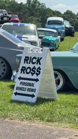 The video shows a sunny outdoor car show. A white A-frame sign with black text that reads "RICK ROSS" and "PERSONAL VEHICLE ROWS" is in the foreground. Behind the sign, several vintage cars are parked on a grassy field under a clear blue sky. The cars appear to be from the 1950s and are of various colors, including teal, light blue, and red. Trees line the background. A person in a green shirt and shorts walks from right to left across the frame.