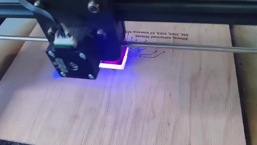 A close-up shot shows a laser engraving machine working on a piece of plywood. The laser head, a black square device with wires connected to it, moves horizontally across the surface of the wood. A bright blue light emanates from the laser head as it engraves a pattern onto the plywood. The engraving appears to be a diagram of a circuit board. Text is also visible on the plywood, reading "3Dnow, enhanced 3Dnow, SSE2, SSE3, 57 Romana Mark" and "2005". The plywood rests on a textured surface, possibly a workbench. The lighting is natural, with the blue light from the laser being the most prominent illumination. The engraving process is methodical, with the laser tracing precise lines on the wood.