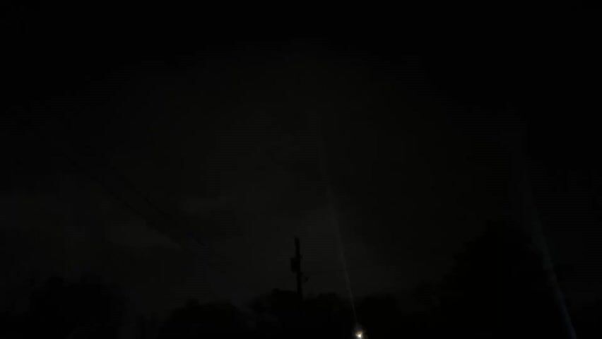 The video depicts a very dark nighttime scene with a single visible star in the sky. In the foreground, there's a silhouetted power pole with a faint glow emanating from a light fixture near its base. The sky appears cloudy. The foreground is also fringed with silhouetted trees and branches. Overhead power lines can be faintly seen.