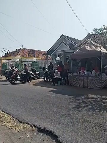 The video shows a street scene, likely in a residential area, with several people and motorcycles. On the right side of the road, there's a temporary setup under a canopy, resembling a small market stall or a community event table. Several individuals are gathered around this area, some appearing to be interacting with each other. There are bottles on the table, and some people are wearing red shirts. To the left, a few motorcycles are parked or in motion on the road. The environment appears to be outdoors during the daytime, with clear skies and sunlight casting shadows. The buildings in the background are modest houses with tiled roofs. Some people are seated, while others are standing. The overall impression is of a casual gathering or a local event taking place on a street.