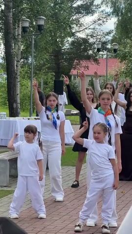 A group of children and young adults, dressed in white, are performing an outdoor dance routine. The front row consists of four children: two young girls and two young boys. They are all wearing white t-shirts and white pants. In the second row, there are three teenage girls. The girl on the left is wearing a white t-shirt and white pants, and has a colorful scarf tied around her neck. The girl in the middle is wearing a black dress. The girl on the right is wearing a white t-shirt and white pants, also with a colorful scarf. Behind them, other participants are visible, also dressed in white or dark clothing.

The scene appears to be a park or an outdoor event space, with trees and greenery in the background. A red-roofed building is visible in the distance, along with some streetlights. There are white tablecloths on tables to the left, suggesting a reception or gathering. The lighting suggests it is daytime, possibly late afternoon. The expressions on the faces of the performers, particularly the younger ones, indicate engagement and enthusiasm for their performance. One of the teenage girls is singing.

There is no visible text in the video.
