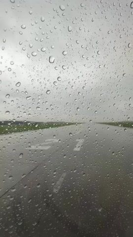 The video shows a close-up view of a window covered in raindrops. Through the raindrops, a runway is visible, with green fields on either side. The sky appears overcast and gray. The perspective suggests the viewer is inside a vehicle, possibly an airplane, as it is on a runway and the raindrops are distorting the view. The video is short and features no discernible actions or interactions.