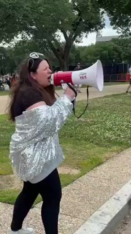 A woman with long dark hair, wearing a black top, black leggings, and a shimmering silver sequined jacket, is actively speaking into a megaphone outdoors. She has sunglasses perched on her head and her mouth is wide open, indicating she is loudly shouting or chanting. The megaphone is red and white, and the words "F*ck ICE" are clearly visible on its side. She is standing on or beside a concrete path, with her body angled to her left.

The setting appears to be a large, open grassy area, likely a park or public space. Numerous green trees form the background, suggesting a natural environment within an urban or suburban area. Several other individuals are visible in the mid-ground and background, some sitting on the grass, others standing further back, hinting at a gathering or event. Black temporary barriers or fencing can be seen in the distance on the right side. The overall lighting suggests it is daytime, possibly overcast or cloudy. The woman's expression and use of the megaphone suggest she is passionate and engaged in a protest or public address in Washington, United States.