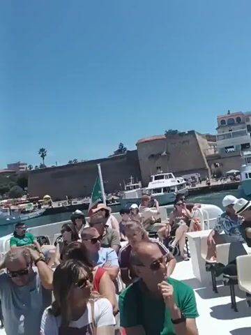 The video shows a group of people on a boat, looking out at a harbor filled with sailboats. The sky is blue and clear, and the sun is shining. The people on the boat are dressed in casual clothing, and some of them are wearing hats to protect themselves from the sun. The sailboats are docked in the harbor, and their sails are furled. The buildings in the background are typical of a coastal town, with terracotta roofs and pastel-colored walls. The water in the harbor is a deep blue, and there are a few other boats in the distance. The overall impression is one of a beautiful, sunny day in a picturesque location.