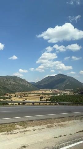 A vertical video shows a view from a moving vehicle looking out at a rural landscape. In the foreground is a paved road with a white line marking the lane. Beyond the road, a grassy shoulder transitions to a field of dry, yellowish crops. A dense line of green trees runs along the edge of the field, separating it from more distant, rolling hills covered in vegetation. Two larger mountains dominate the background, with a valley between them. The sky is a bright blue, dotted with fluffy white clouds. The overall impression is of a sunny day in the countryside.