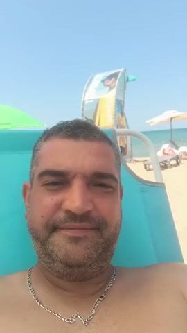 A man with short, dark hair and a beard is lying on a beach chair. He is wearing a silver necklace. The man is looking at the camera. The background is a beach with umbrellas and lounge chairs. The sky is blue. The video is recorded in portrait mode.