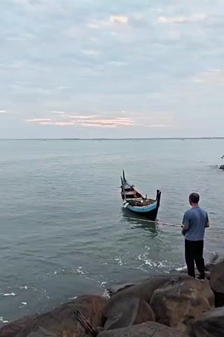 The video shows a man standing on a rocky shoreline, looking out at a small, traditional-style boat floating in the ocean. The boat is primarily dark in color with a blue stripe along the top edge. The man is wearing a light blue shirt and dark pants. The sky is overcast with faint sunset colors near the horizon. The ocean is relatively calm, with small waves lapping at the shore. Several other small boats are visible in the distance.