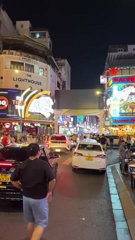 This media file captures a vibrant street scene at night, likely in Thủ Đức, Vietnam. The primary subjects are people and vehicles navigating a busy urban environment. The setting is a street lined with buildings adorned with numerous neon signs, indicating a commercial and entertainment district.

The activity taking place is street life at night. People are seen walking, interacting, and sitting at outdoor establishments. Vehicles, including cars and taxis, are present on the road, suggesting a flow of traffic. One taxi, with "TAXI" clearly visible on its roof, is a prominent object in the foreground.

Notable details include the time of day, which is clearly night, with artificial lighting dominating the scene. The weather appears to be clear, as there are no visible signs of rain or inclement conditions. The abundance of neon signs, some with animal motifs like a tiger, and the architecture of the buildings provide strong location cues. The overall atmosphere appears lively and bustling, with a sense of energy associated with a popular nightlife area.

Visible text includes "THE LIGHTHOUSE" on a building, "TIGER" next to a neon tiger graphic, and "TAXI" on a vehicle's roof. On a distant archway, "PHO DI BO BUI VIEN" is visible, a common street name associated with tourist and entertainment areas in Vietnam. There are also numerous other signs with text in Vietnamese, contributing to the energetic and culturally specific ambiance.