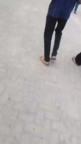 The video shows a ground-level view of a public square or marketplace with a gray brick or cobblestone surface. Several people are walking around in different directions. Some are wearing casual clothes such as shorts and t-shirts, while others are wearing more formal attire.  In the background, there's a large metal structure providing shade, likely a market or transportation hub covering. Buildings and an elevated road can be seen in the far background under an overcast sky. The video is somewhat shaky, suggesting it was filmed handheld while walking.