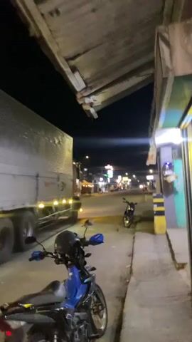 The video captures a nighttime scene on a street. In the foreground, two motorcycles are parked on a sidewalk. One is blue and black, and the other is black. A large white box truck is moving away from the camera on the street, its taillights and side marker lights illuminated. In the background, the street continues with traffic, streetlights, and buildings visible. The overall atmosphere is that of a typical urban or suburban street at night.