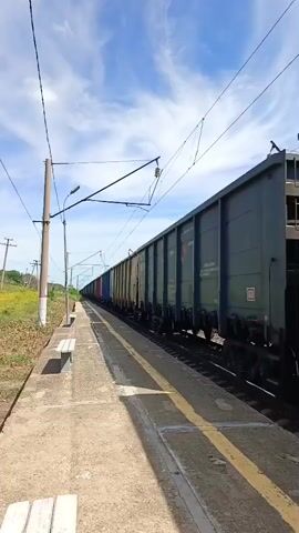 A long train of freight cars passes a deserted train station platform. The train cars are of various colors, including green, blue, red, and beige. The platform is made of concrete and has a single bench. Power lines run overhead, supported by poles. The sky is blue with scattered white clouds. The scene is captured from a low angle, with the train dominating the right side of the frame. The train appears to be moving from left to right in the video.