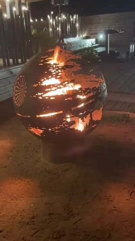 The video shows a fire pit shaped like the Death Star from Star Wars. The fire pit is round and made of metal with cutouts that allow the flames to be visible. The fire is burning brightly inside the Death Star. The fire pit is located outdoors on a sandy or dirt ground. In the background, there is a wooden deck with some lights strung up. There are also some plants and furniture visible on the deck. The overall scene is one of a cozy evening outdoors with a fire.