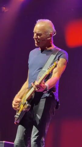 A video shows a man playing a bass guitar on a stage. He is wearing a blue t-shirt and dark pants. He has a headset microphone. The stage is lit with purple and red lights. The man appears to be singing and playing.