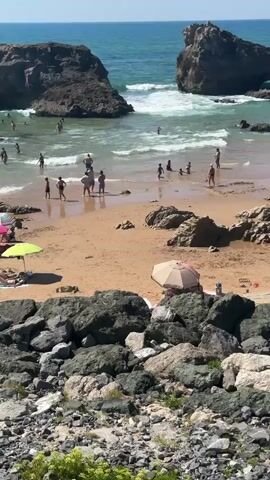 The video shows a bustling beach scene on a sunny day. The ocean is a vibrant turquoise with white-capped waves gently rolling towards the shore. Large, dark rocks jut out from the water and are scattered along the sandy beach. Numerous people are enjoying the water, swimming, playing, and wading. On the sand, several beachgoers are relaxing under colorful umbrellas, including pink, green, and blue ones. Some people are sunbathing, while others are sitting or standing near the water's edge. The overall impression is one of a lively and popular summer beach destination.