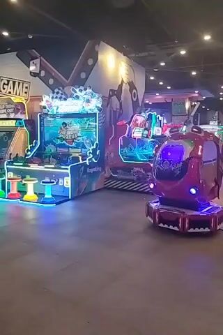 The video shows an indoor arcade with multiple game machines. In the foreground, on the left, is a pink and red kiddie ride shaped like a car, with a young child inside. To the right of the kiddie ride is a large, brightly lit arcade game machine featuring a racing theme, with multiple seats and controls. Further to the right, another arcade game machine with a racing theme is visible, also brightly lit with neon lights. In the background, more arcade games are visible, along with overhead lighting and decorative elements on the walls. Several people are present, including adults and children, some playing games and others observing. The floor is tiled. The overall atmosphere appears lively and colorful.