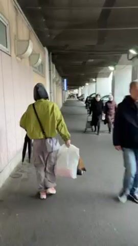 A person wearing a green jacket, baggy pants, and pink sneakers walks away from the camera, carrying a plastic bag and a brown paper bag. The person also carries an umbrella on their shoulder. They walk on a concrete walkway covered by a concrete overhang. The area is also occupied by parked cars and several other people, some of whom are pulling shopping carts. The area looks like a parking lot or a loading area beside a building.