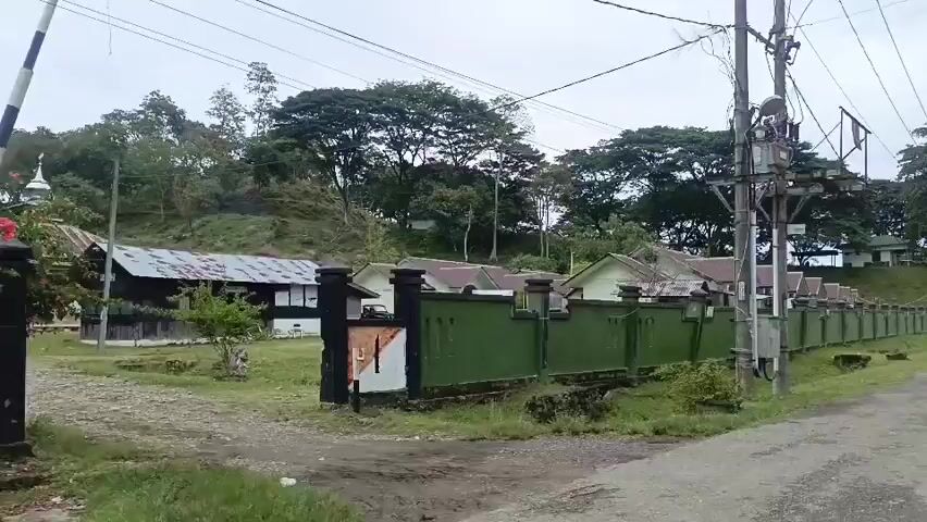 A video captures a roadside view of a village or compound. The foreground features a paved road and a green wall that runs along the side of the road. Behind the wall, there are several houses with light-colored walls and darker roofs, some with visible antennas. A mosque with a dome and a minaret is also visible within the compound. The area is surrounded by lush green vegetation and trees. The sky is overcast. The camera is moving, creating a panning effect that reveals more of the scene. There are no people visible in the video. The overall impression is a quiet, rural setting.
