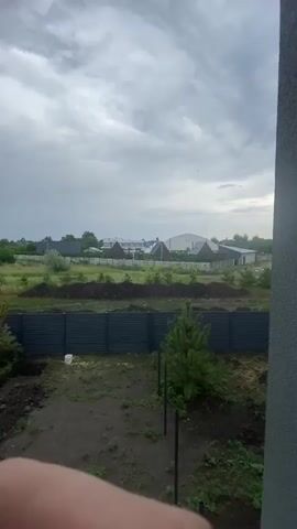 The video captures an outdoor scene, likely a residential area under construction or undergoing landscaping. A dark, corrugated metal fence runs horizontally across the foreground, with a small evergreen tree to the right. Beyond the fence, a patch of uneven ground is visible, with piles of dark soil suggesting recent excavation or earthmoving. Further back, a low concrete block wall separates this area from a grassy field dotted with young trees and saplings. In the background, several buildings are visible, including houses with distinct triangular roofs, a larger white structure resembling a warehouse or sports facility, and other less distinct buildings. The sky is overcast with grey clouds. The overall impression is one of an undeveloped or developing residential property.