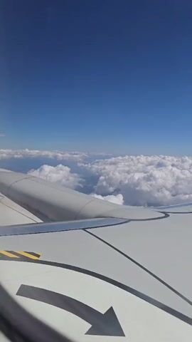 The video shows a view from an airplane window looking out at the sky and clouds. The wing of the airplane is visible in the foreground. The sky is blue, and there are many white, fluffy clouds below the airplane. The view is serene and peaceful.