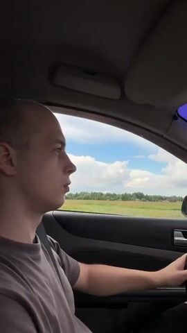 A person is driving a car. The passenger side window is visible, showing a green landscape with trees and a blue sky with clouds. The driver is looking forward. The car interior is dark. A white building with blue lettering is visible through the window. The sound of a car engine and music is heard.
