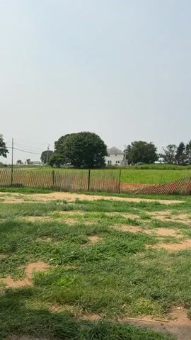 The video shows a rural landscape on a hazy day. In the foreground, there is patchy green grass and exposed dirt. A wooden fence, partially covered with orange netting, runs horizontally across the middle of the frame. Beyond the fence, there is a field of green crops. In the background, several houses are visible, along with clusters of trees. The sky is a pale, washed-out blue. The camera appears to be panning slowly to the right.