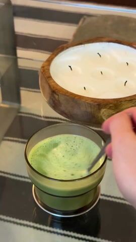 A person’s hand stirs a glass of green liquid with a metal spoon on a glass-topped table with a black and beige striped rug underneath.  A large, round wooden bowl holds a white wax candle with several black wicks.