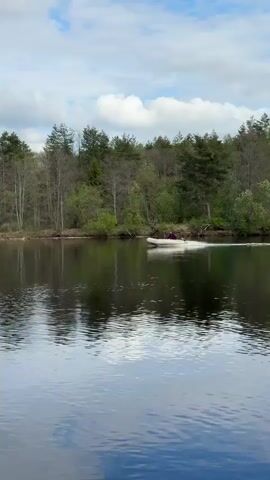 The video shows a lake on a partly cloudy day. The lake water is calm with rippling reflections of the sky and trees on the opposite shore. The shore is lined with trees, some of which still have bare branches, while others are green. In the middle of the lake, a white inflatable boat with a person in it is moving from right to left. The boat is kicking up a spray of water behind it as it moves. The person in the boat is wearing a dark-colored top. The boat continues moving across the lake.