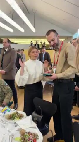 The video shows a woman in a white blouse and black skirt standing in a room with several people and tables. She gestures with her hands, then embraces another woman wearing a dark blazer. There are tables with plates of food, and a man in a khaki shirt with a badge around his neck stands nearby. The room appears to be at a conference or meeting, with many people dressed in business attire visible in the background. The lighting is bright from overhead lights.
