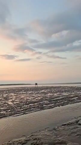 A man stands with his arms crossed looking out at the water during sunset. He is standing next to a partially ruined brick structure with graffiti on the wall. The mudflats stretch out before the water, and the sky is filled with soft clouds.