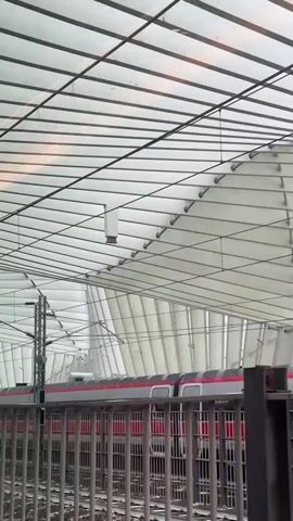 A train with red and white stripes is moving to the right behind a metal fence. The train is inside a modern train station with a translucent roof made of white panels and metal beams. There are electrical wires and poles visible above the train. The camera appears to be looking through the fence from ground level.