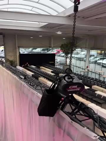The video shows a large indoor space, seemingly set up for an event. In the foreground, a chain hoist labeled "CM Lodestar" hangs from the ceiling, attached to a metal truss structure. A black bag is also attached to the truss. Below the truss, a white curtain hangs down. Rows of chairs and cushioned benches are arranged on a platform, facing a stage area. Beyond the seating, there are tables and a bar setup. Large windows line the back of the space, offering a view of a parking lot filled with cars. Red lights illuminate the lower level. The camera pans across the scene, also revealing a second-level balcony area with desks, equipment, and people working. The overall environment appears to be a showroom or event space being prepared for an audience.