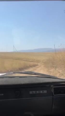 The video provides a view from inside a Lada car as it travels down a dirt road through a dry, grassy field. The sky is clear and blue. There are a few utility poles visible in the distance. The dashboard of the car is visible in the foreground. The car appears to be moving at a moderate speed.