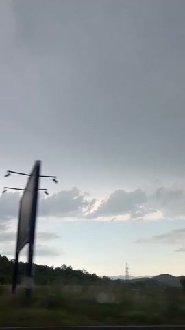 This video captures a scenic view from a moving vehicle, likely a car, as it travels through a landscape under a cloudy sky. The dominant subject is the expansive sky, filled with varying shades of gray and lighter patches indicating breaks in the clouds. Below the sky, a line of trees and rolling hills forms the horizon.

Notable details include a power transmission tower visible on the left side of the frame, suggesting the presence of infrastructure within the natural setting. The greenery of the trees and the contours of the hills indicate a rural or semi-rural location. The overall lighting suggests either early morning or late afternoon, with the diffused light characteristic of overcast conditions. The movement of the vehicle is evident from the slight blur of the trees and foreground, implying the footage was recorded while in transit.

The weather appears to be cloudy, possibly leading up to or following rain, given the slightly muted colors and the mood conveyed by the sky. There are no visible people or discernible text in the video. The scene evokes a sense of journey and contemplation, with the vastness of the sky and the natural landscape contributing to a peaceful, albeit somber, atmosphere. The location context of Gospoština, Montenegro, places this scene in a region known for its natural beauty, often characterized by mountainous terrain and coastal influences.
