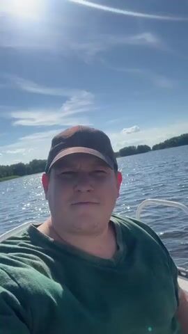 A man wearing a green t-shirt and a brown baseball cap is sitting on a boat. He is looking at the camera and smiling. The boat is on a lake, and the sun is shining. There are trees in the background. The water is choppy and there are some clouds in the sky. The camera is moving slightly, as if the boat is in motion.
