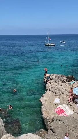 This is a vertical video showing a vibrant scene at a rocky coastline, likely near Palermo, Italy. The bright, clear blue sky and the sparkling turquoise water suggest a sunny day with excellent weather. The sun is high, indicating it's midday or early afternoon.

The main subject is a person in black shorts who is captured mid-jump, leaping from a rocky outcrop into the clear water below. The splash and whitewater indicate they have just entered the sea. Nearby, on the same rocky ledge, another person stands watching. Several other people are visible in the water, swimming and enjoying the sea. Some are further out, appearing to be swimming or relaxing.

In the background, the deep blue sea stretches out to the horizon. A white sailboat with a red flag is anchored or sailing in the distance, adding to the idyllic coastal atmosphere. Another smaller boat is also visible. The rocky shore is dotted with people sunbathing on towels. A pink and white striped towel is prominently placed on the rocks.

The overall mood is one of leisure and summer fun. The clear, inviting water and the act of cliff jumping convey a sense of adventure and enjoyment of a beautiful summer day. The presence of boats suggests a popular spot for recreation and tourism. There is no visible text in the video.