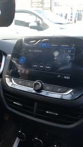 A car sits at a traffic intersection with several other cars. A building on the corner houses various businesses. The dashboard of the car is visible, showing a radio station playing and the time 10:03.