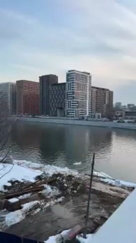 The video shows a view of a river with modern buildings on the opposite bank. The buildings are various shades of gray, brown, and white, with different patterns on their facades. The river has a slight ripple to it, and there are some patches of ice and snow along the riverbank. The foreground has some snowy patches mixed with dirt and debris. A few bare trees are also visible near the river's edge. The sky is overcast with a blue hue.
