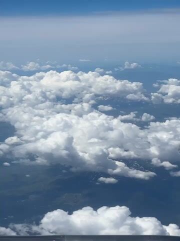 This is a vertical, aerial view from an airplane window, looking down at a vast expanse of clouds and the landscape below. The top of the frame shows a clear blue sky that transitions to a lighter haze near the horizon. Below the sky, a thick layer of white, puffy cumulus clouds dominates the scene, casting shadows on the terrain beneath. Patches of green and brown land are visible through gaps in the clouds, with hints of snow-capped mountains in the distance. The lower part of the frame includes a blurry horizontal band, which appears to be the edge of the airplane's window or wing. There are no people or discernable objects in the foreground or midground, and no actions or interactions are taking place. The overall impression is one of serene, expansive natural beauty from a high altitude.