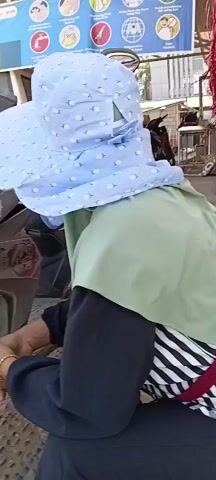 A person wearing a light blue, patterned headscarf and a light green hijab is visible from behind. They are also wearing a black garment with a striped fabric peeking out at the bottom and a red strap across their chest. The person appears to be interacting with something near a dark-colored motorcycle, indicated by their hands being clasped in front of them. The background shows a paved area with hexagonal tiles, a fence, buildings with pink walls, and several parked motorcycles. The lighting suggests it is daytime.